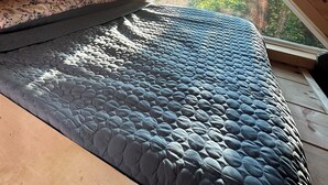 Un dormitorio rústico en el ático con techo inclinado, cama con colchón azul texturizado y vistas a árboles por la ventana.