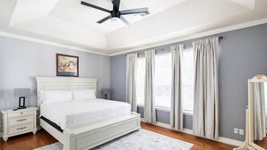 Ein Schlafzimmer mit einem weißen Bett, einem Deckenventilator, einem Nachttisch mit Lampe und einem gerahmten Bild an der Wand.