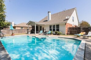 A rectangular swimming pool with a covered patio area, lounge chairs, and a wooden fence in the background.