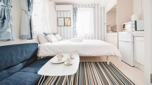 A compact hotel room with a bed, a sofa, a small table with tea set, a kitchenette, and striped curtains.