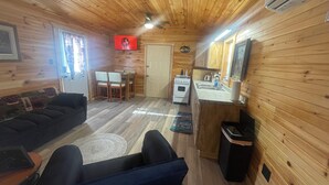 A cozy cabin interior with a wooden ceiling, a dining area with chairs, a sofa, a kitchen with a stove, and a TV mounted on the wall. - Deer Run Cabin (Licking)