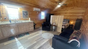 A cozy cabin interior with a wooden ceiling, a kitchen area with a sink and cabinets, a seating area with a couch and a chair, and a bed with a headboard. - Deer Run Cabin (Licking)