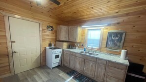 A compact kitchen with wooden walls, a white stove, a sink, and a window with blinds. - Deer Run Cabin (Licking)