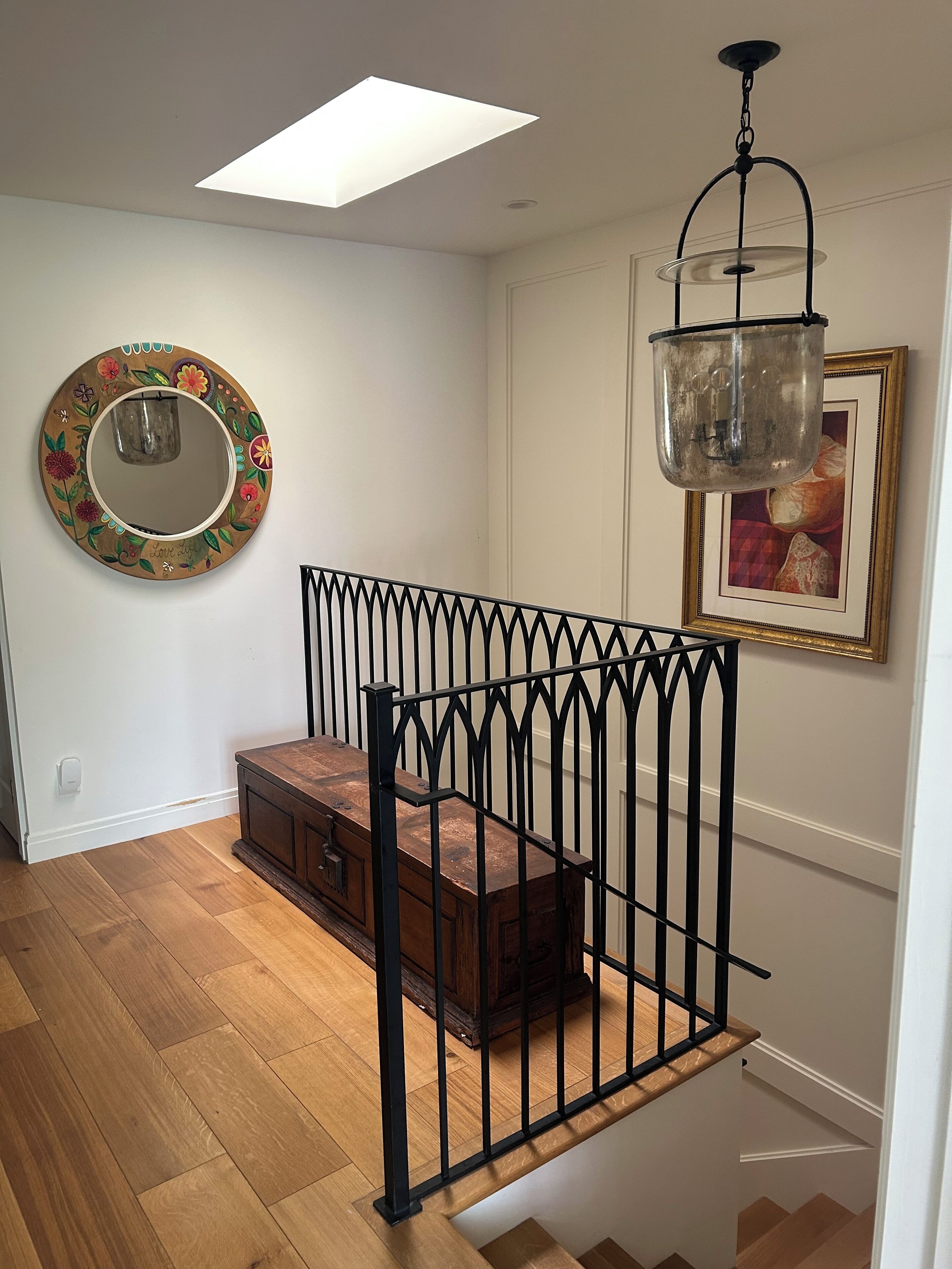 A staircase with a black metal railing, a wooden bench, a round mirror with a floral design, and a framed picture on the wall.
