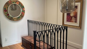 Una escalera con barandilla de metal negro, un banco de madera, un espejo redondo con diseño floral y un cuadro en la pared.