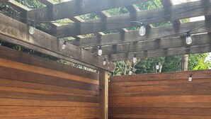 Una pérgola de madera con luces colgantes, una mesa blanca con cubierta estampada y asientos acolchados azules con cojines oscuros.