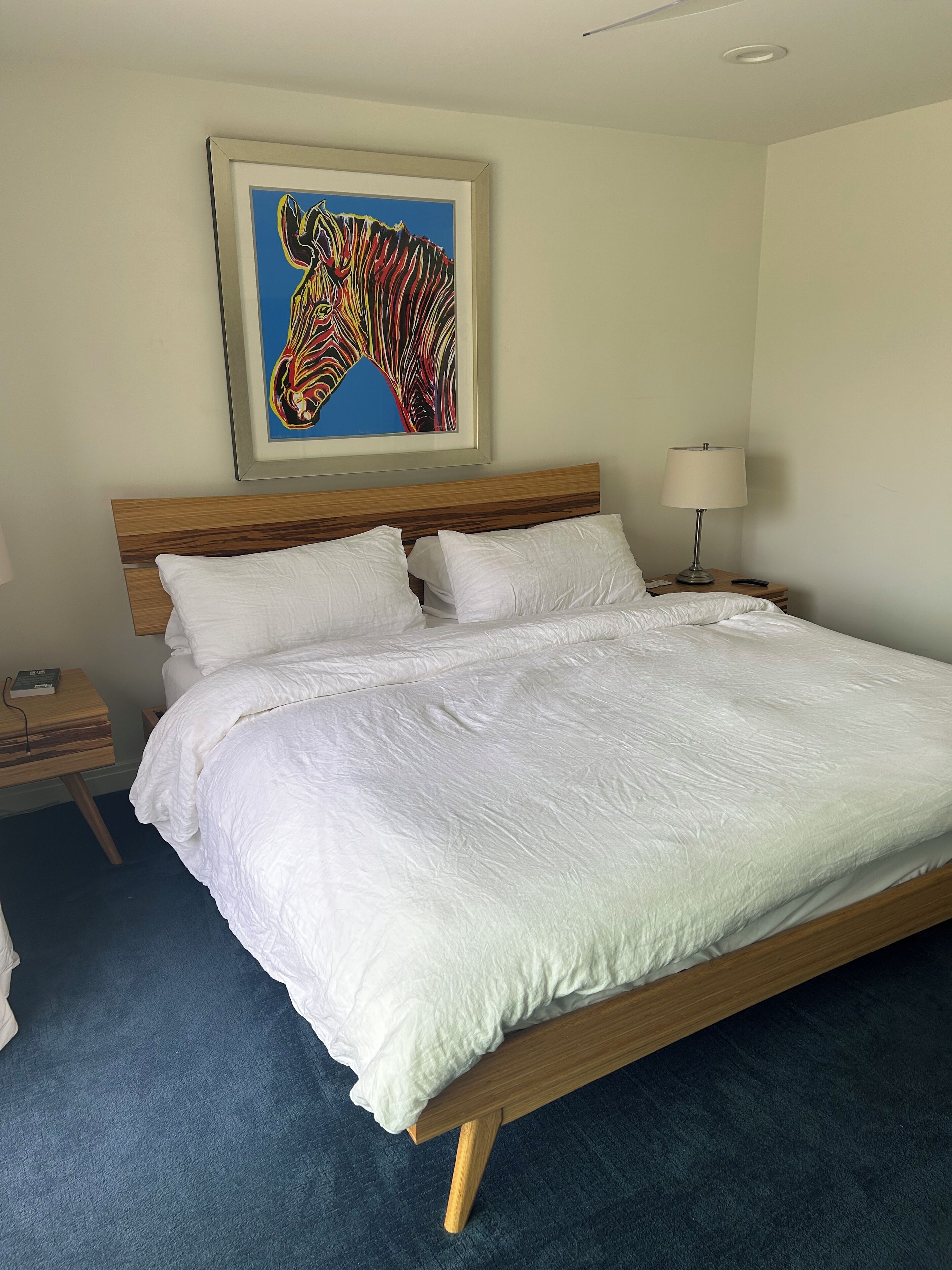 A neatly made bed with white bedding, a wooden headboard, a framed abstract painting above, a bedside table with a lamp, and a dark blue carpet.