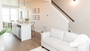 Una cocina moderna con una isla central, un área de comedor con una mesa y sillas blancas, y una escalera que sube a un nivel superior.