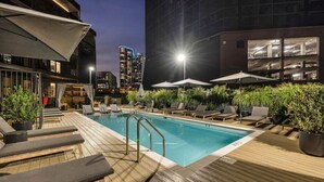 Zona de piscina en la azotea con sillones de descanso y sombrillas, rodeada de vegetación y edificios modernos al fondo.