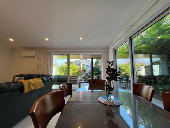 Una sala de estar moderna con una mesa de comedor de cristal, sofás de cuero y un amplio ventanal corredero que da a un balcón con vegetación.