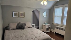 Un dormitorio con cama, ventilador de techo, ventana con persianas, radiador y una mesita de noche con caja de pañuelos.