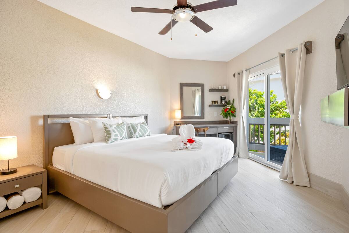A hotel room with a large bed, a ceiling fan, a nightstand with a lamp, a window with curtains, and a view of greenery outside.