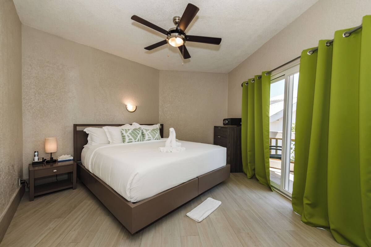 A hotel room with a bed, a nightstand, a lamp, a ceiling fan, and a view of the outdoors through a window with green curtains.