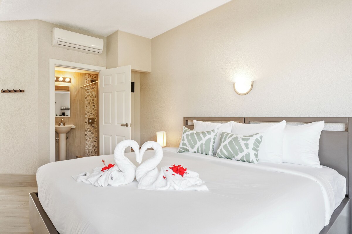 A hotel room with a bed, a wall-mounted air conditioner, a bathroom with a sink and mirror, and a nightstand with a lamp.
