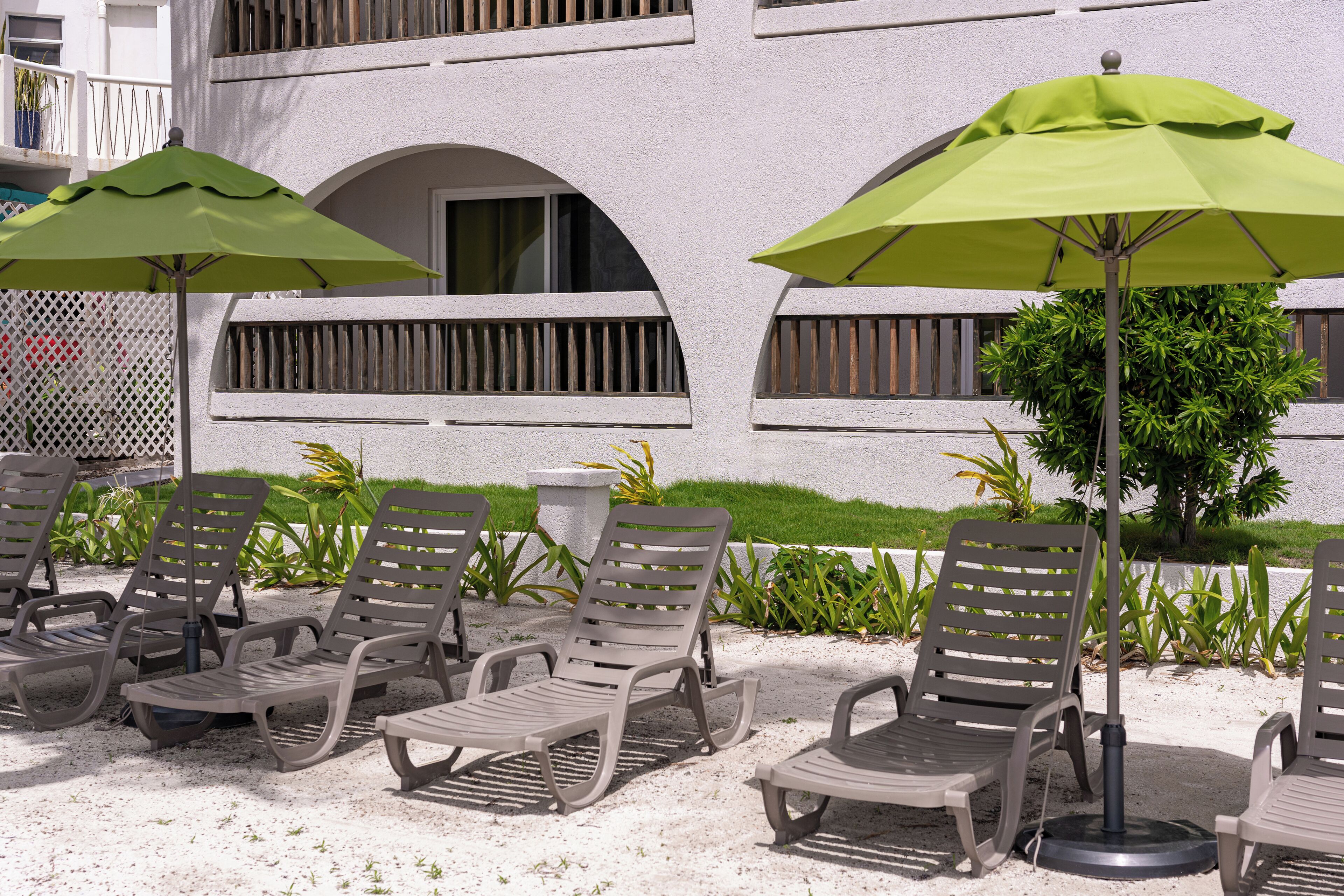 Outdoor lounge chairs with umbrellas, a white building with balconies, and a small tree.