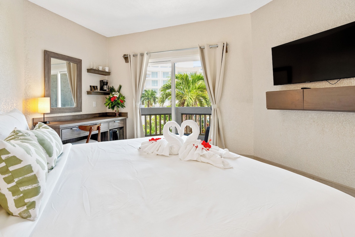 A hotel room with a bed, a desk, a chair, a TV mounted on the wall, and a window with curtains.