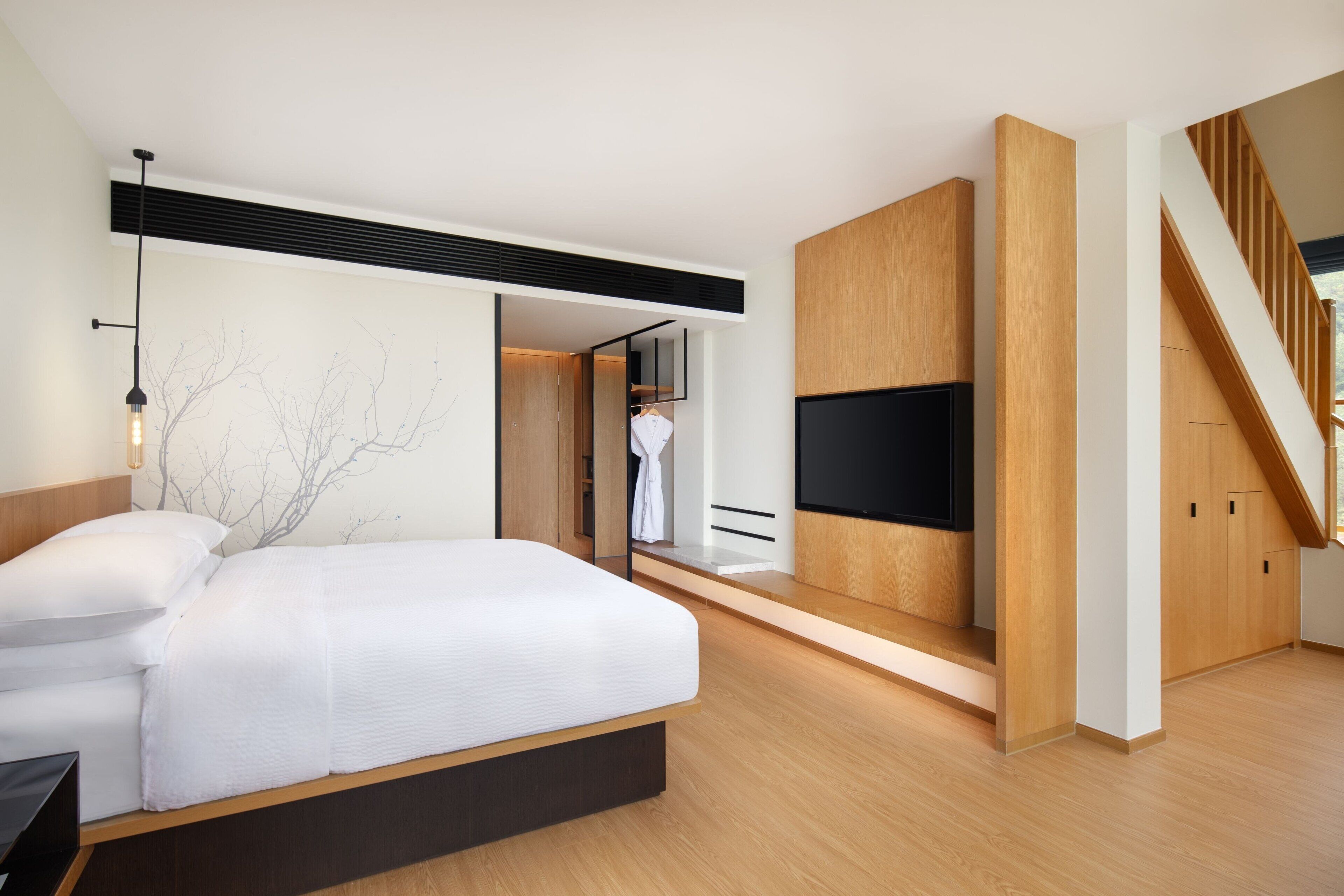 A modern bedroom with a large bed, built-in TV, wooden flooring, and a staircase.