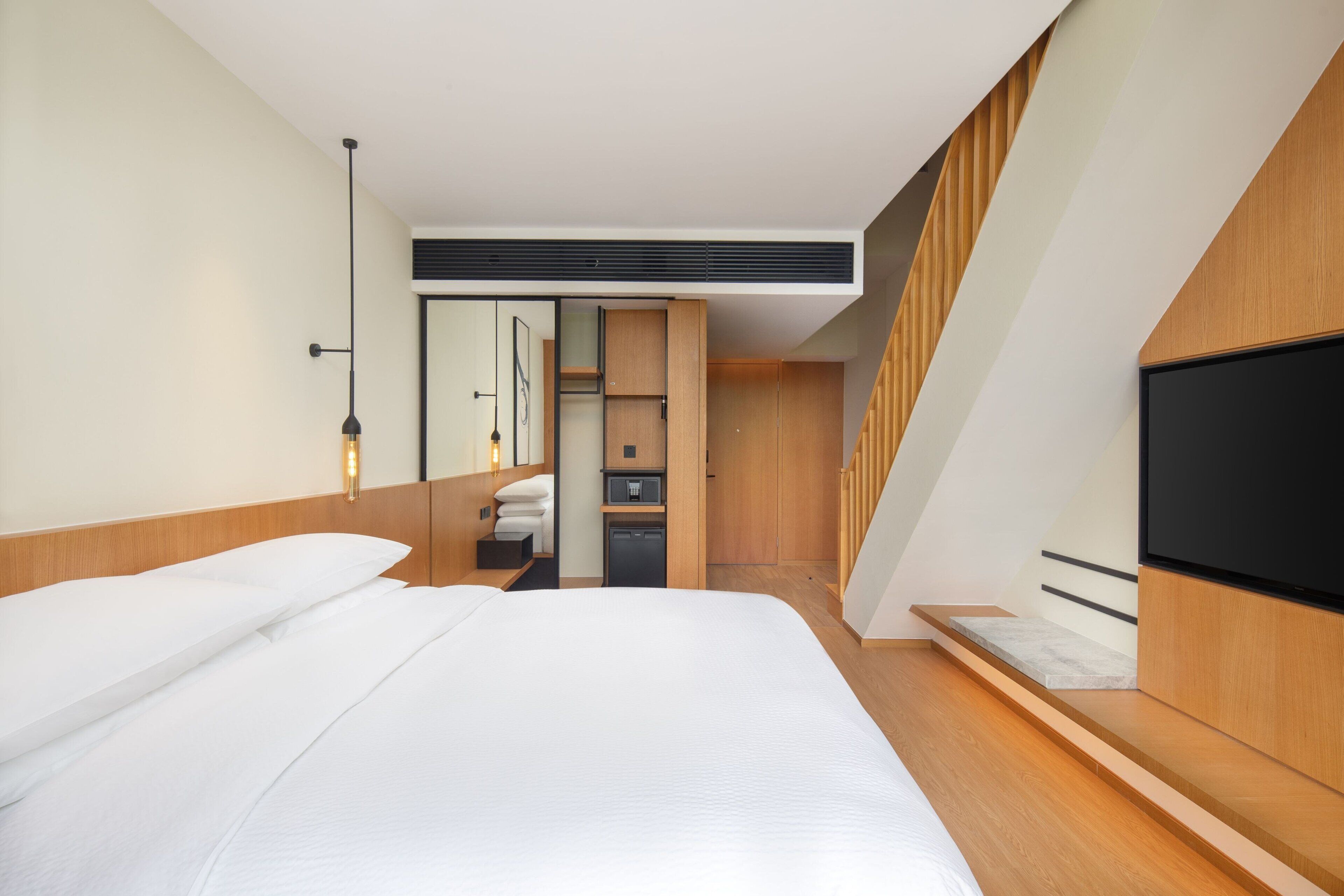 Una habitación de hotel moderna con una cama grande, un televisor de pantalla plana, un armario empotrado y un pequeño rincón de descanso con una banca.