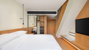 Una habitación de hotel moderna con una cama grande, un televisor de pantalla plana, un armario empotrado y un pequeño rincón de descanso con una banca.
