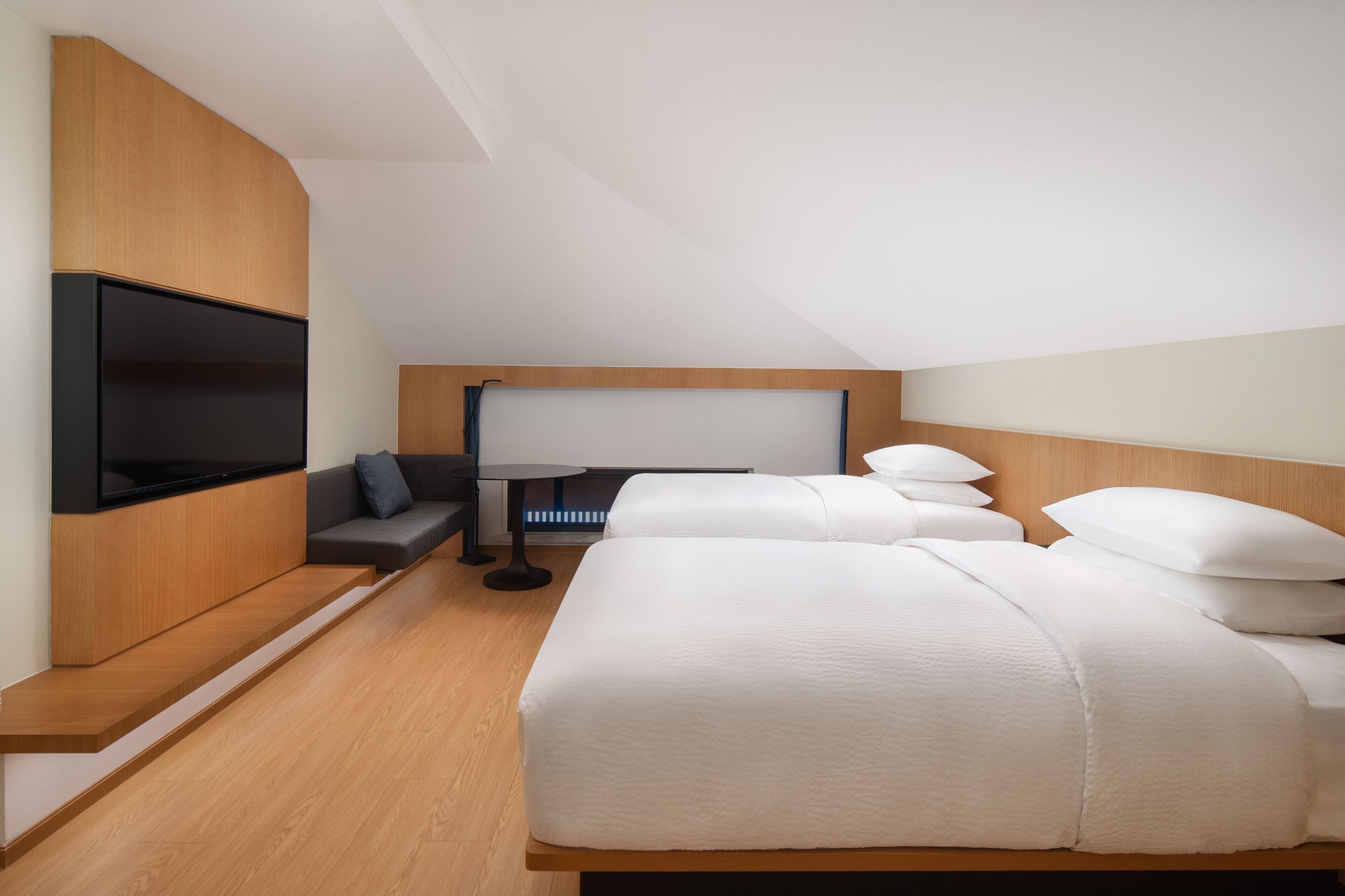 A modern hotel room with a large bed, a flat-screen TV mounted on a wooden panel, a small table, and a sofa.