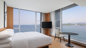 Una habitación de hotel moderna con una cama grande, un televisor, una mesita y una silla. La habitación tiene ventanas amplias con vistas al mar y a las montañas.