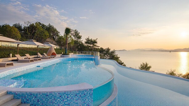 Una piscina infinita curva con vistas al mar, rodeada de sillones y sombrillas.