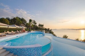 Una piscina infinita curva con vistas al mar, rodeada de sillones y sombrillas.