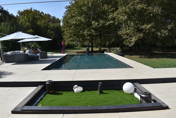 Area piscina esterna moderna con una piscina rettangolare, una zona relax con ombrellone bianco e un prato curato.