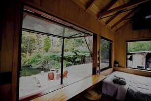 A cabin with a large window, a bed, a table, and a chair. - Charming Tiny House Escape
 (Bald Knob)