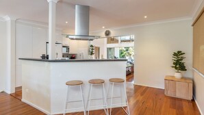 Una cocina moderna con una isla central, armarios blancos y una campana extractora de acero inoxidable. Hay tres taburetes en la isla y una planta en maceta sobre un mueble de madera.