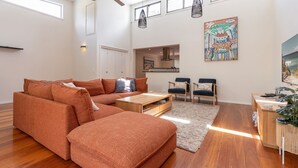 Una sala de estar moderna con un sofá modular, una mesa de centro y dos sillones. Se observa una cocina al fondo, un cuadro en la pared y una planta al lado.
