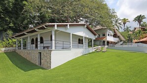Un bâtiment blanc doté d’un patio couvert et d’un socle en pierre, entouré d’une pelouse bien entretenue et de palmiers.