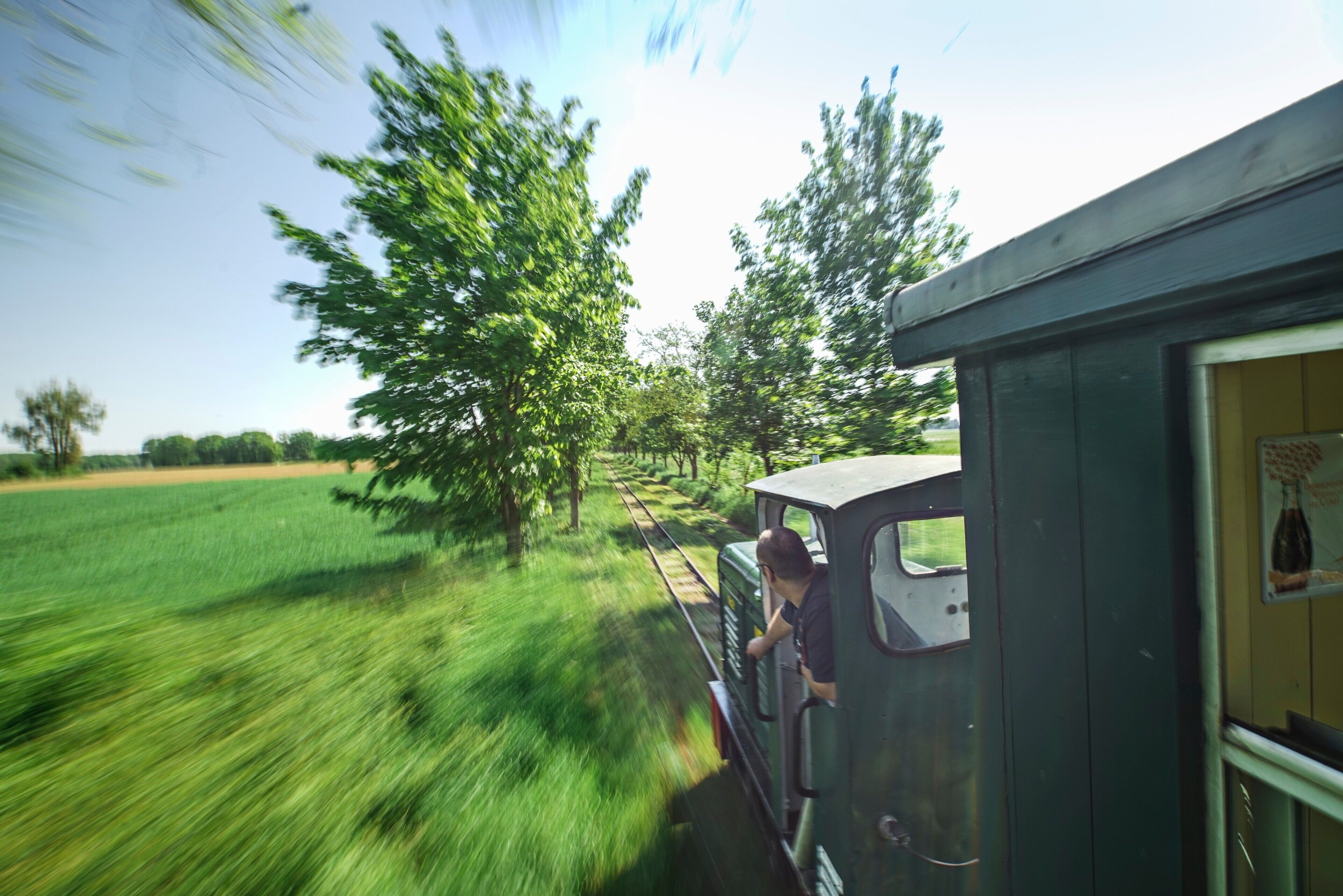 A vintage train with a person standing at the front, traveling through a rural area with green fields and trees.