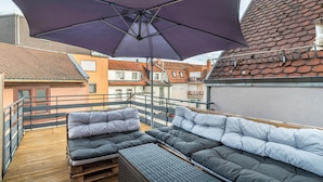 Eine Dachterrasse mit großem Sonnenschirm, Holzdeck und einer Couch mit Kissen.