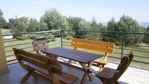 En terrasse med træmøbler, gelænder i metal og udsigt til grøn natur og bygninger.