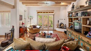 A spacious living room with a fireplace, wooden shelves, and a television. - Quail Run 640 | Kiva Fireplace | Private Patio | 2-Car Garage | Golf Course | Pool & Hot Tub (Santa Fe)