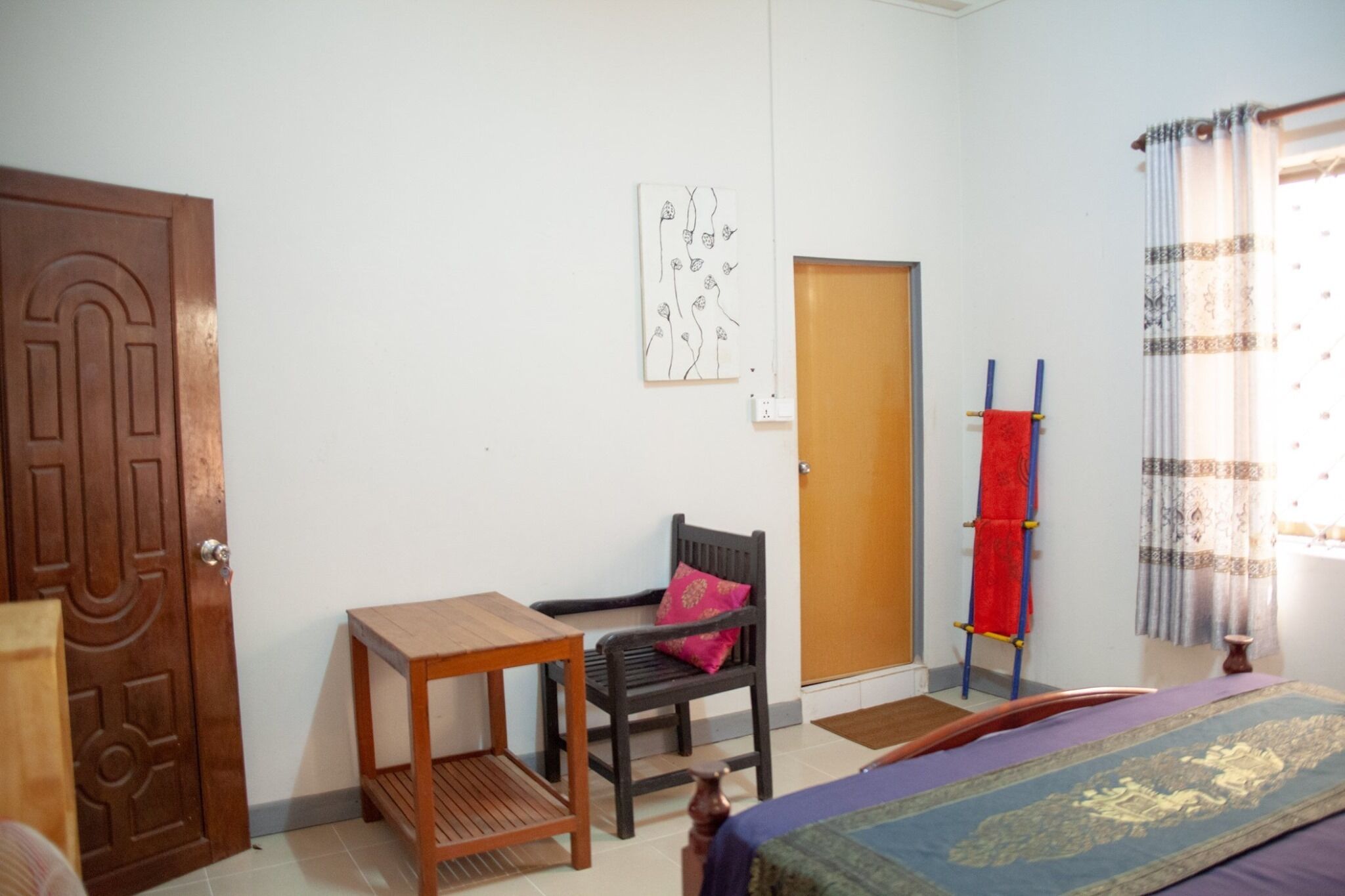 A room with a wooden door, a small wooden table, a black chair with a red cushion, a bed with a patterned bedspread, a wall-mounted artwork, a wooden door, a red towel rack, and a window with patterned curtains.