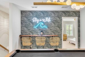 A pool table with three wicker chairs, a neon sign reading "Stay a While," and a room with a bed and dresser in the background.