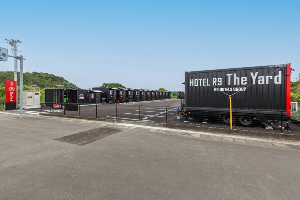 Ein großer schwarzer Container mit der Aufschrift "HOTEL R9 The Yard" und "R9 HOTELS GROUP", abgestellt auf einer Straße, im Hintergrund blauer Himmel und viel Grün.