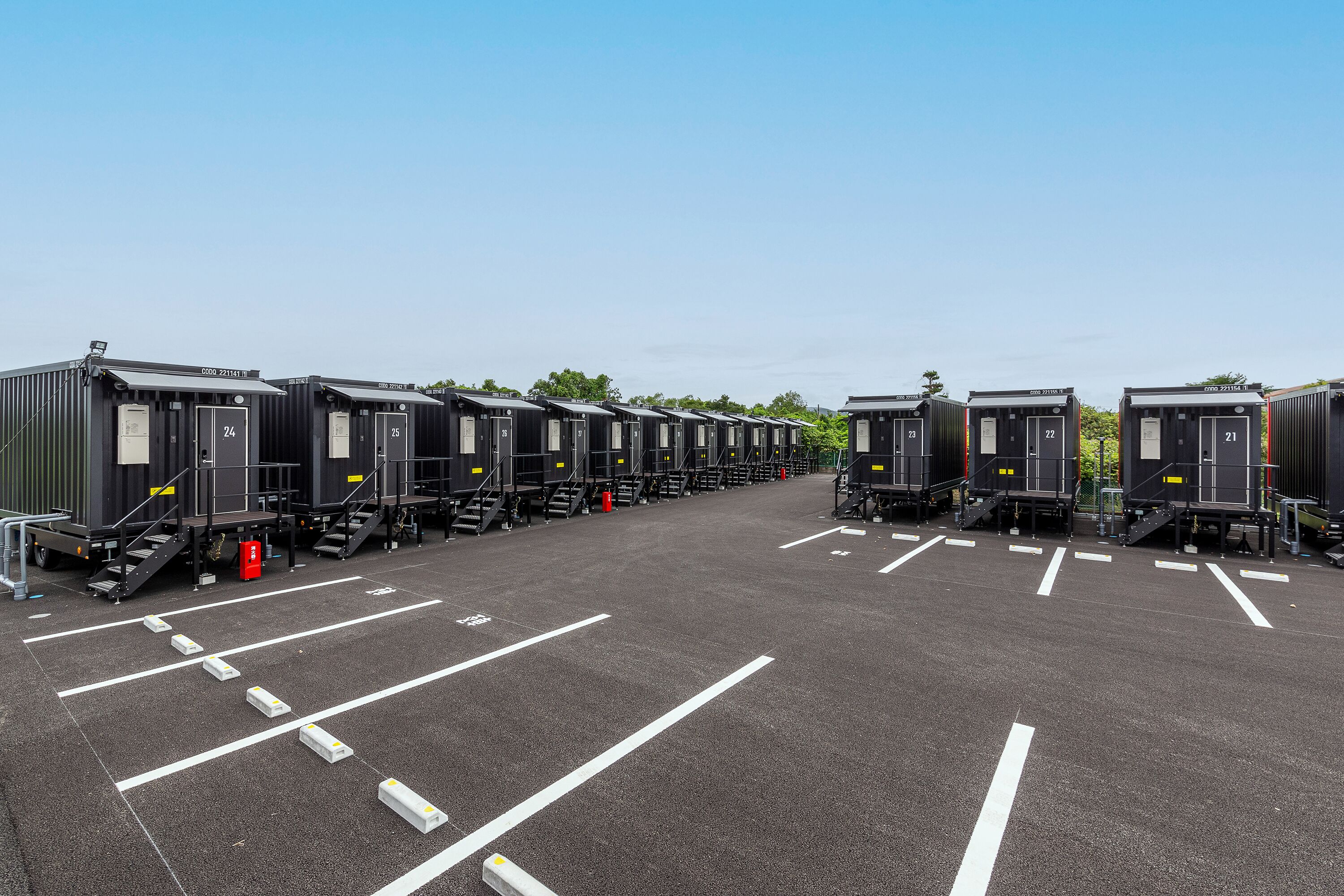 A row of portable cabins with numbered doors, arranged in a parking lot with marked parking spaces.