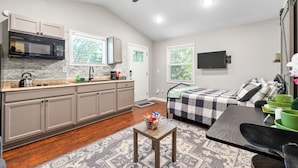 Un espace de vie compact comprenant un coin cuisine, un lit recouvert d’une courtepointe à carreaux, une table à manger avec des chaises et un ventilateur de plafond.