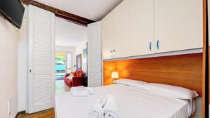 A hotel room with a bed, white curtains, a wooden headboard, a TV mounted on the wall, and a door leading to another room.
