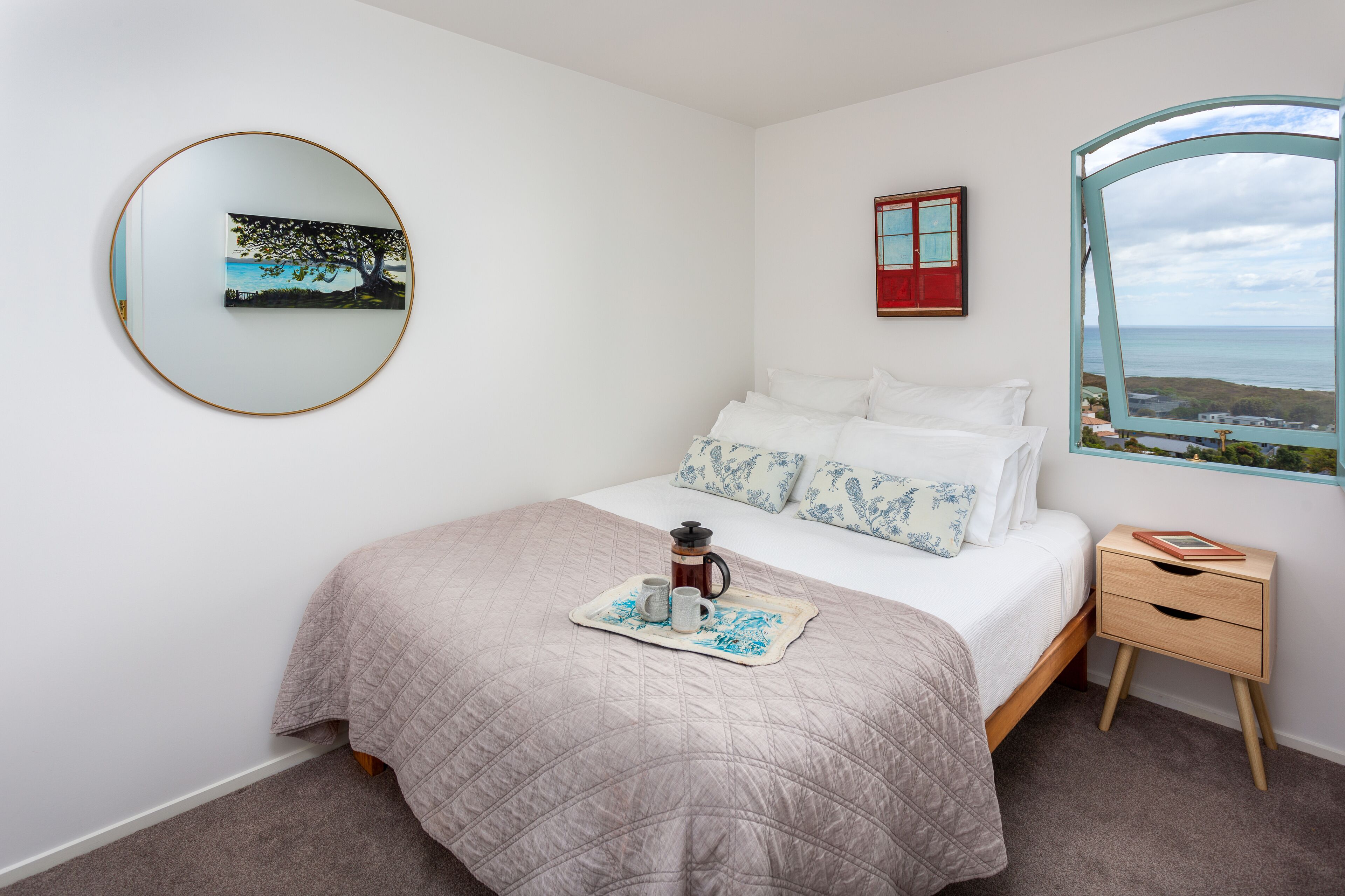 A bedroom with a bed, a bedside table, a coffee maker, and a view of the sea.