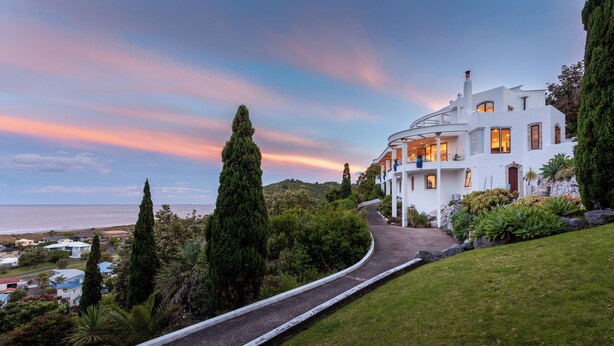 A white house with a curved driveway, surrounded by greenery and overlooking a coastal area.