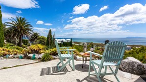 Two light blue Adirondack chairs facing a table with a drink and a plate of food, overlooking a scenic view of the sea and distant land.
