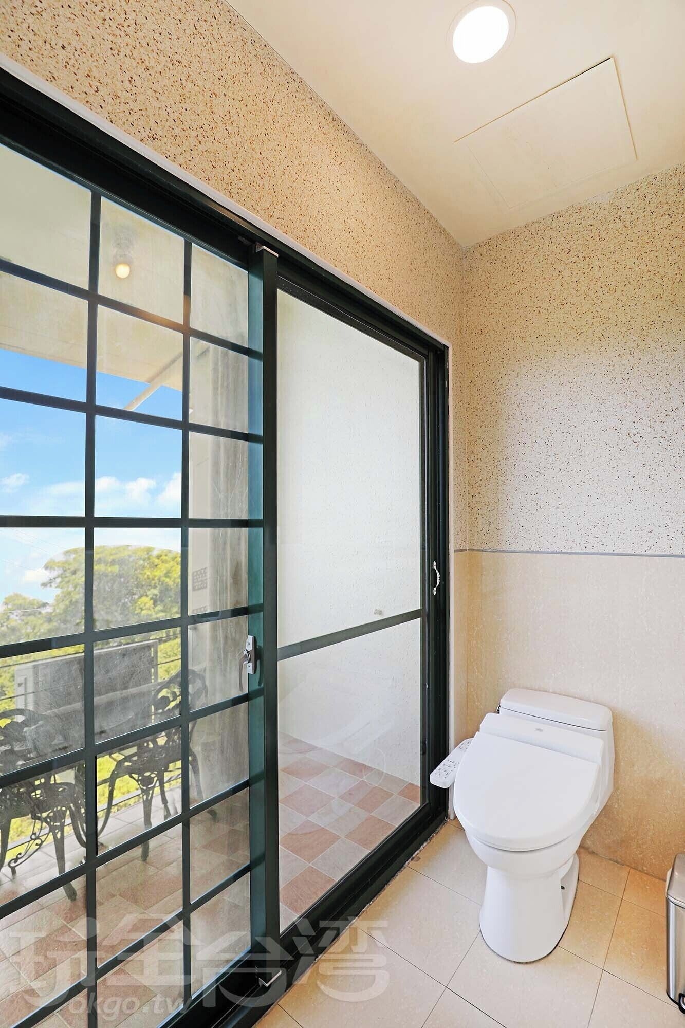 Un baño con inodoro, una puerta de vidrio que da a un espacio exterior con mesa y sillas, y suelo de baldosas.