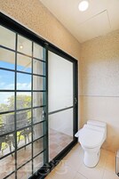 Un baño con inodoro, una puerta de vidrio que da a un espacio exterior con mesa y sillas, y suelo de baldosas.