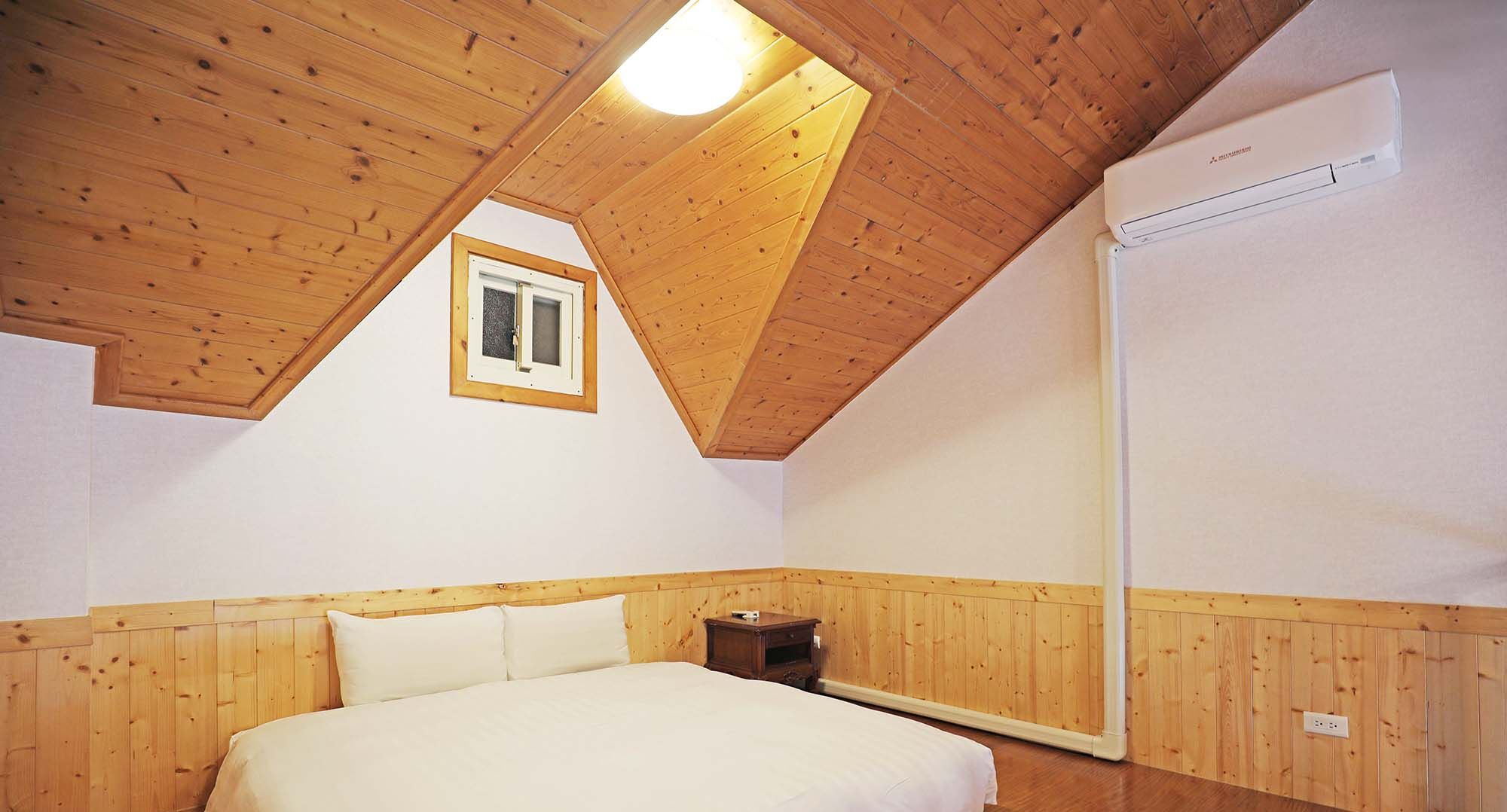 A bedroom with a sloped wooden ceiling, a single bed, a small bedside table, and a wall-mounted air conditioner.