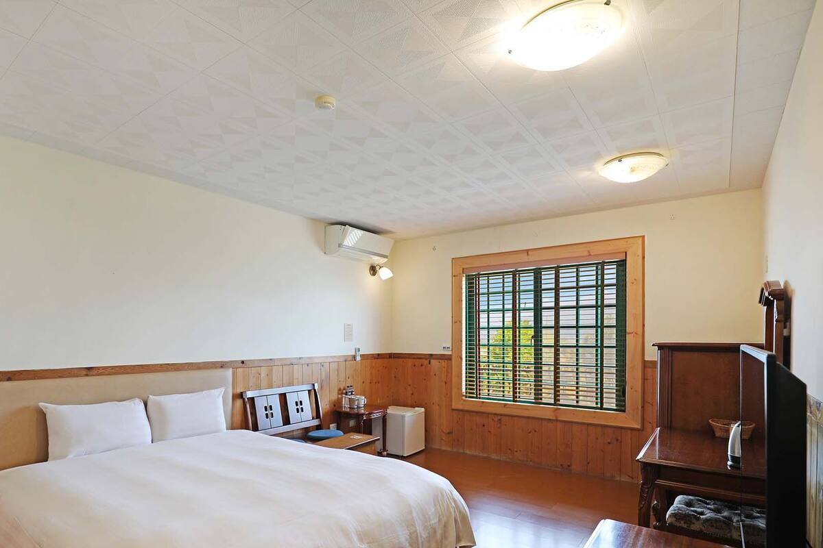 A bedroom with a bed, a small table, a cabinet, and a window with blinds.