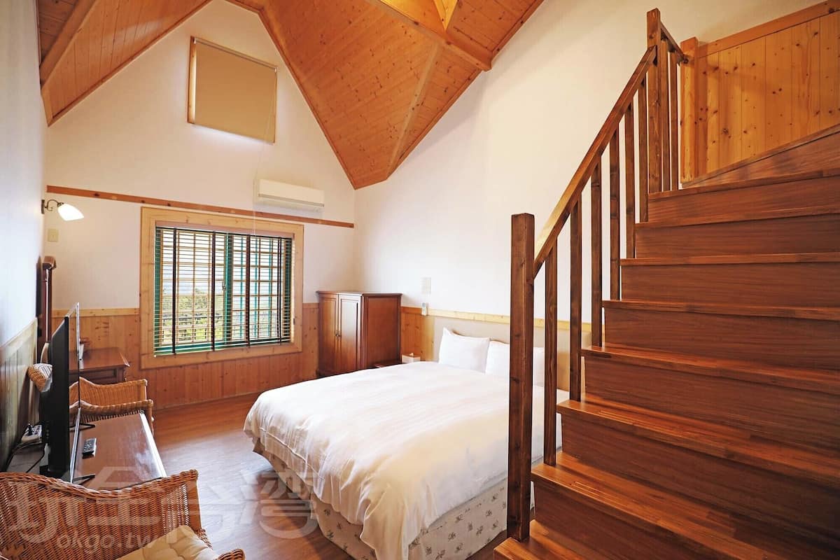 A bedroom with a wooden staircase, a large window with blinds, a bed, a desk, and a chair.